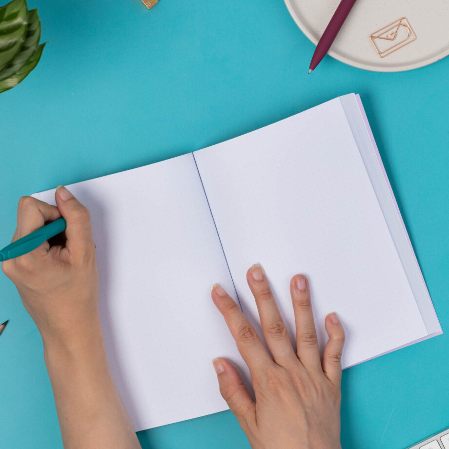 An open A5 Dot Grid notebook, showing the double spread of the dot-grid pages. The right hand is holding the notebook open at the middle, whilst the right hand holding a teal pen is writing in the top left corner of the page. The book is on a teal desk with a white keyboard and other pens around it.
