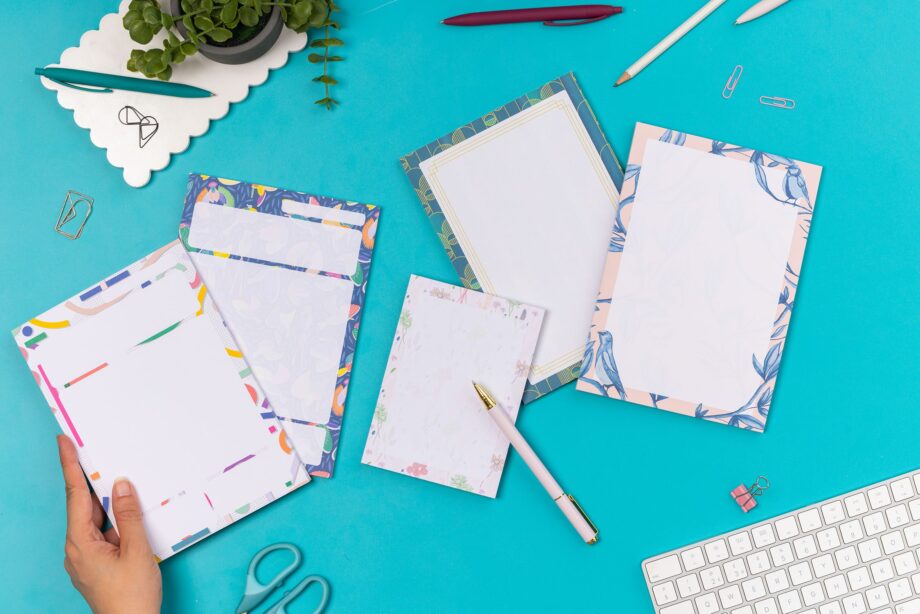 4 A5 notepads (Cutouts, Paradise, Deco Delights and Brighton Birds) are flat in a row with the A6 Home Grown pad, on a teal desk with pens, paperclips and a white keyboard around them. There is a hand holding the cutouts pad in the bottom left of the image.