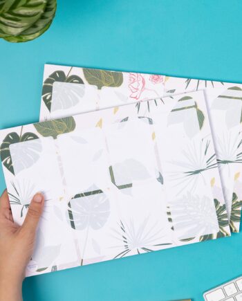 A4 Planner pad with a blush pink background and tropical and monstera leaf details. There are 8 semi-transparent rectangles on the planner, plus a small row on the top right, possibly for a date. A hand is holding the planner pad on the left, with an identical pad underneath. They are on a teal desk with a white keyboard and green plant to the corners of the image.
