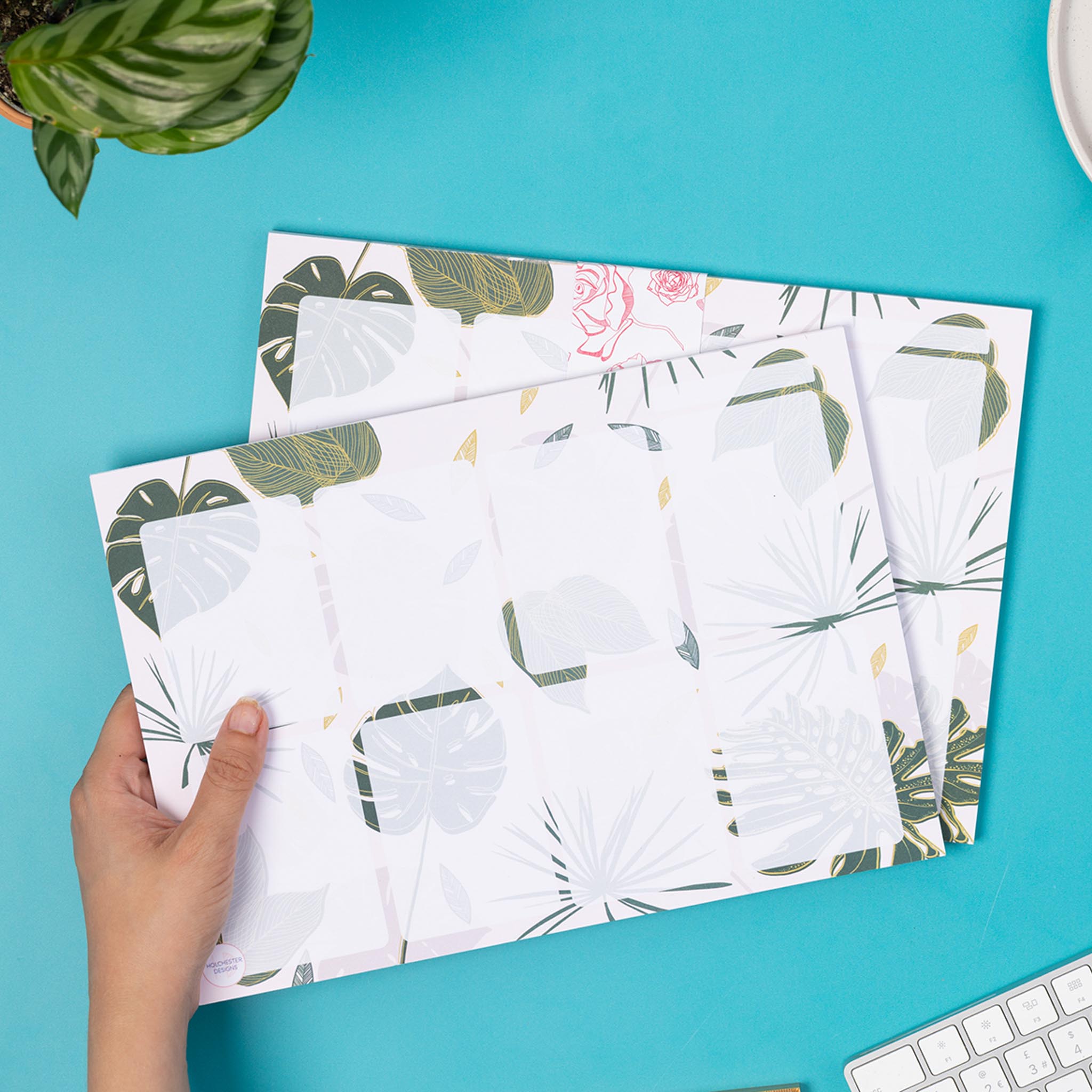 A4 Planner pad with a blush pink background and tropical and monstera leaf details. There are 8 semi-transparent rectangles on the planner, plus a small row on the top right, possibly for a date. A hand is holding the planner pad on the left, with an identical pad underneath. They are on a teal desk with a white keyboard and green plant to the corners of the image.