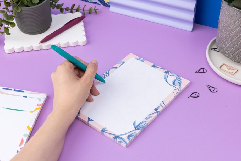 Brighton Birds A5 Notepad - Blush pink background with shades of blue bird and plant hand-painted motifs - is on a lilac desk, next to a white keyboard and scattered paperclips. There is a hand to its left holding a teal pen ready to write on the pad.