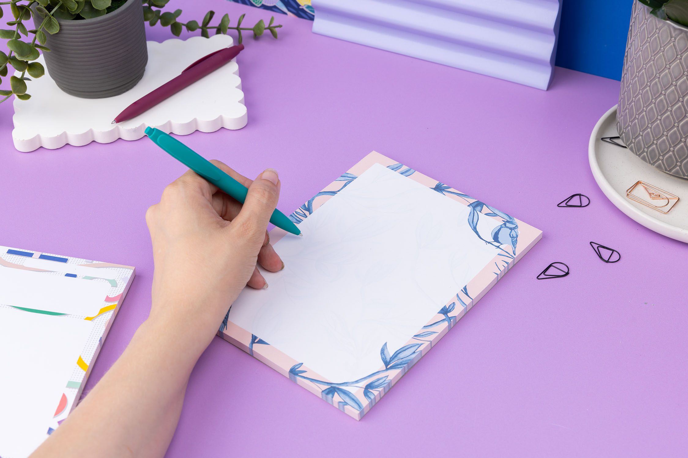 Brighton Birds A5 Notepad - Blush pink background with shades of blue bird and plant hand-painted motifs - is on a lilac desk, next to a white keyboard and scattered paperclips. There is a hand to its left holding a teal pen ready to write on the pad.