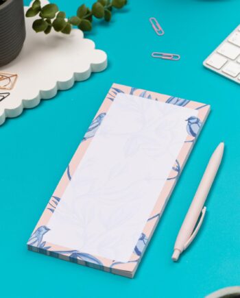 Brighton Birds List pad - Blush pink background with shades of blue bird and plant hand-painted motifs - is on a teal desk, next to a white pen, white keyboard and scattered paperclips.