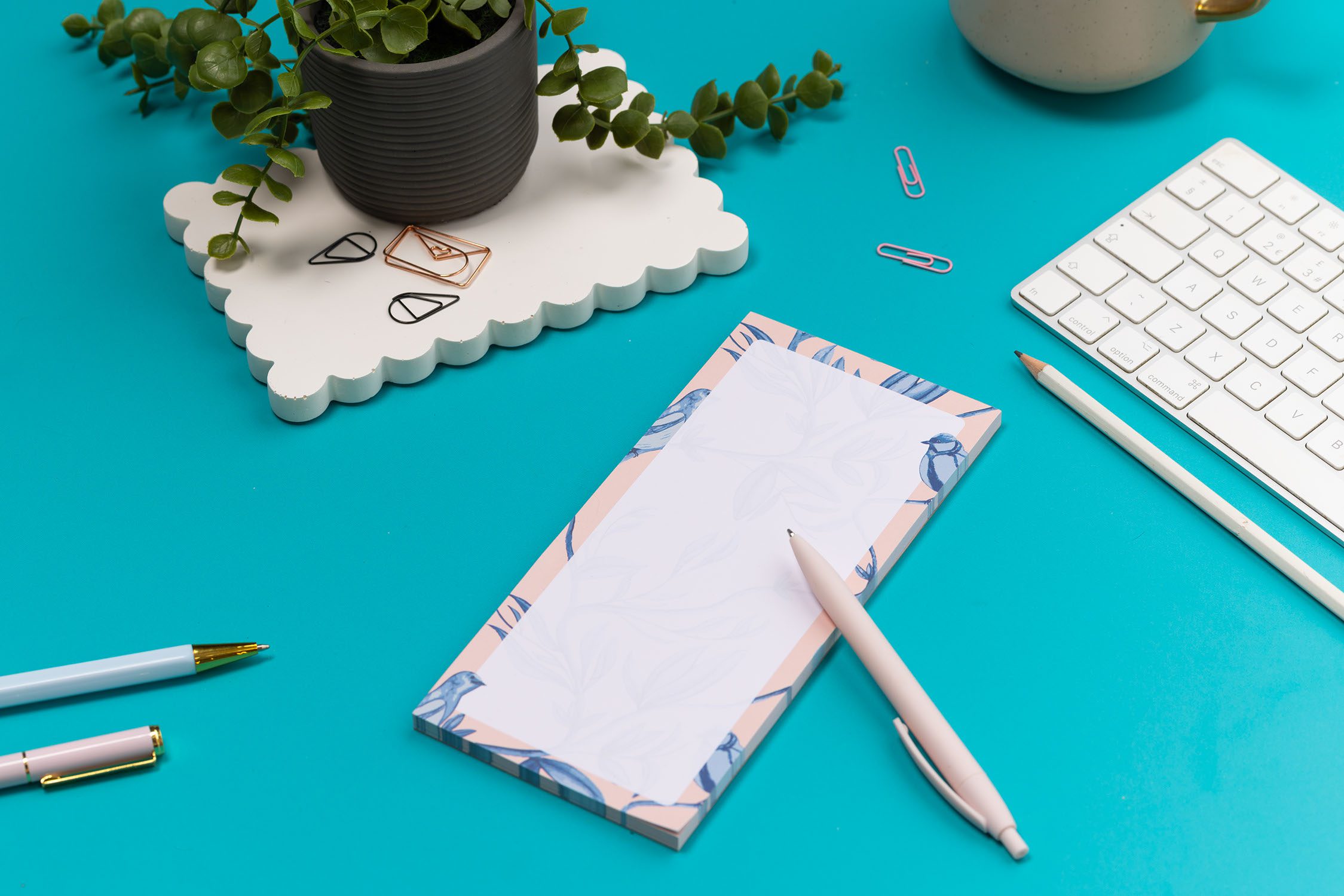 Brighton Birds List pad - Blush pink background with shades of blue bird and plant hand-painted motifs - is on a teal desk, next to a white pen, white keyboard and scattered paperclips.