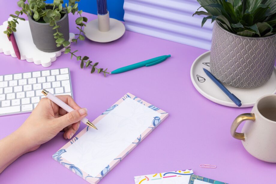 Brighton Birds List pad - Blush pink background with shades of blue bird and plant hand-painted motifs - is on a lilac desk, next to a white pen, white keyboard and scattered paperclips. A hand is on the left of the image, holding a pen to write on the notepad.
