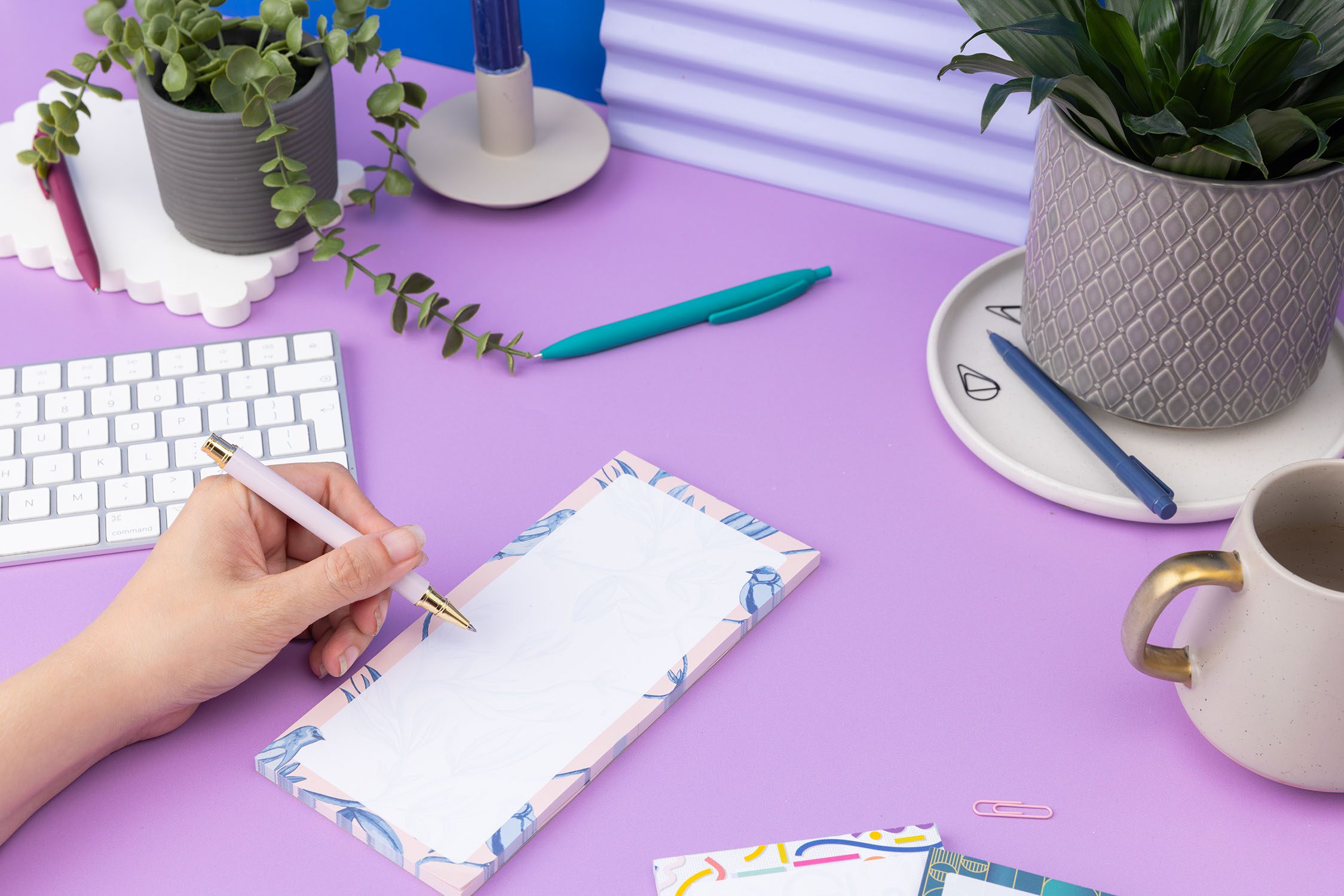 Brighton Birds List pad - Blush pink background with shades of blue bird and plant hand-painted motifs - is on a lilac desk, next to a white pen, white keyboard and scattered paperclips. A hand is on the left of the image, holding a pen to write on the notepad.