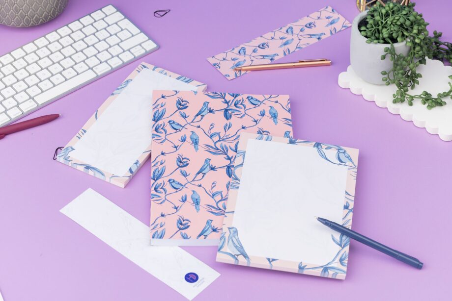 Brighton Birds Full Colleciton - B5 Notebook, List pad, A5 Notepad and 2 double-sided bookmarks (one side up each) - is on a lilac desk, with a white keyboard and pens and paperclips scattered around them.
