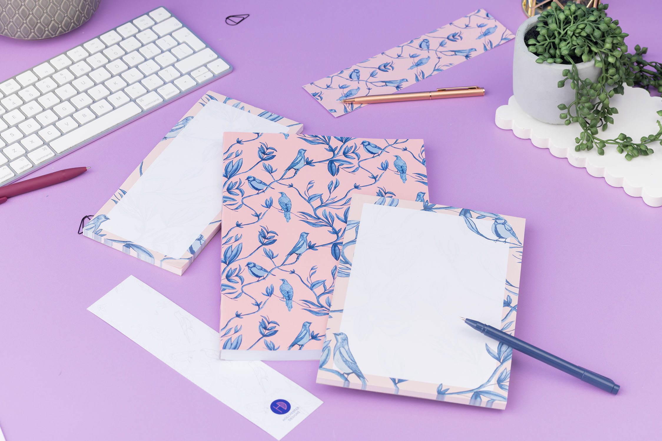 Brighton Birds Full Colleciton - B5 Notebook, List pad, A5 Notepad and 2 double-sided bookmarks (one side up each) - is on a lilac desk, with a white keyboard and pens and paperclips scattered around them.