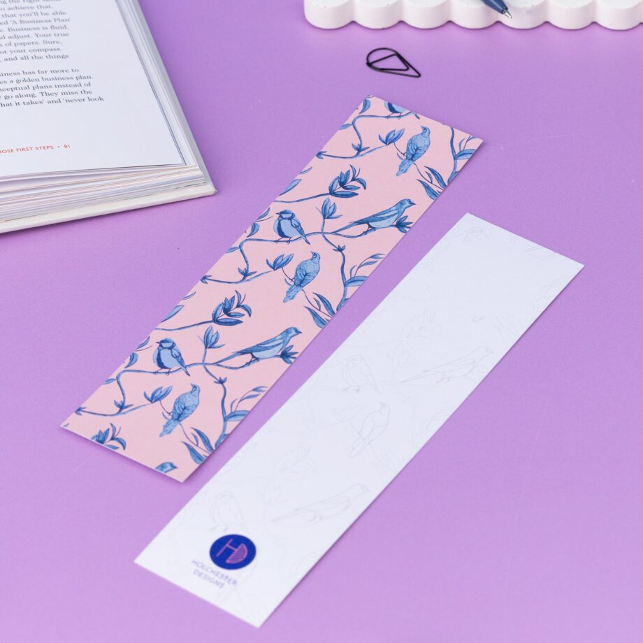 Two of the Brighton Birds double-sided bookmarks, on a lilac desk with a book open to the top left corner. One side features the full-colour patterned side of the Brighton Birds pattern with the blush pink background and shades of blue branch, leaf and bird hand-painted motifs. The other side is the light-grey line drawing.