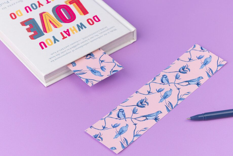 Brighton Birds double-sided bookmark is full-colour pattern side up on a lilac desk. To its left is a closed book which has the same bookmark sticking out the top.
