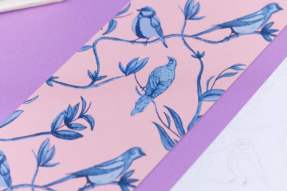 Close up of the full-colour patterned side of the Brighton Birds bookmark with a blush pink background and the blue leaves, branches and birds illustrations. Next to is is an out of focus corner of the bookmark's other side. They are on a lilac desk with an open book in the top left corner.