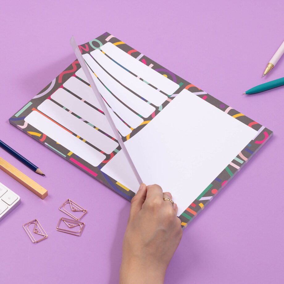 A4 Planner pad with black background, and multicoloured abstract shapes across it. This pad has 7 rectangular boxes in rows on the left of the pad, with one large white rectangular box on the right half. It is on a lilac desk with a hand lifting the top sheet as if to tear it off.