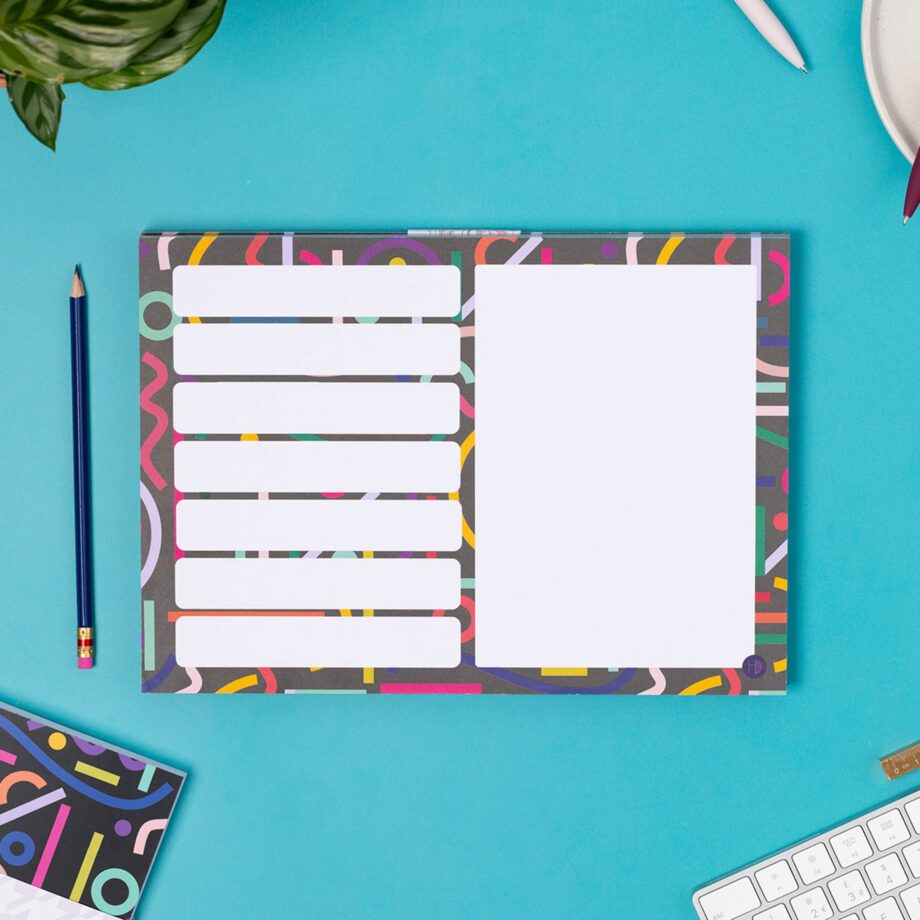 A4 Planner pad with black background, and multicoloured abstract shapes across it. This pad has 7 rectangular boxes in rows on the left of the pad, with one large white rectangular box on the right half. It is on a teal desk with the matching notebook to it's left.