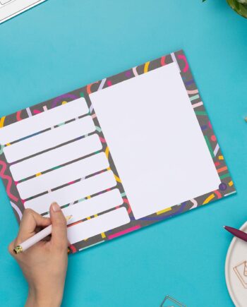 A4 Planner pad with black background, and multicoloured abstract shapes across it. This pad has 7 rectangular boxes in rows on the left of the pad, with one large white rectangular box on the right half. It is on a teal desk with a hand holding a pen to write on it, to the left.