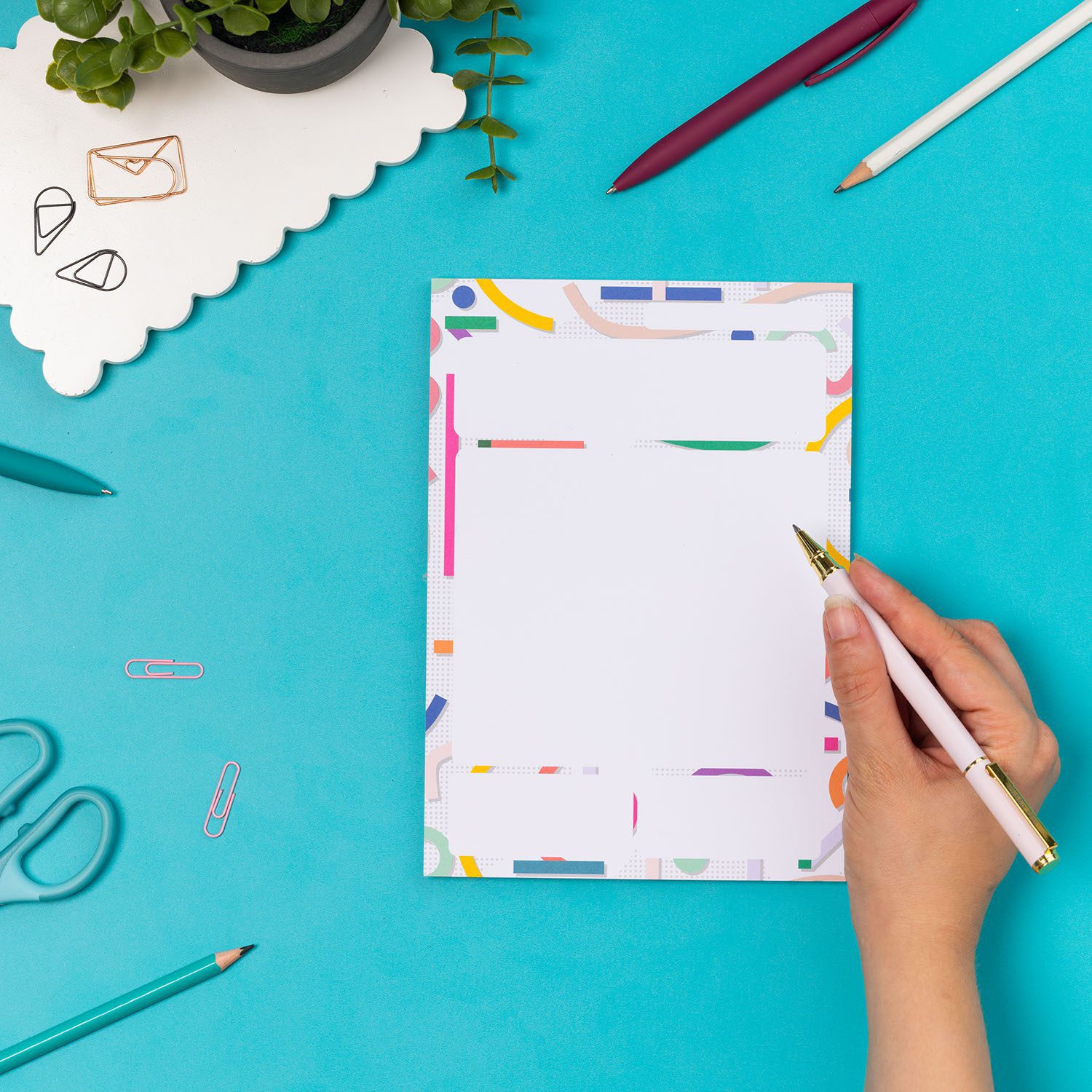 Cutouts A5 Daily Planner Pad on a teal desk, with a hand to the right holding a white pen, and with a small plant and other pens around it.