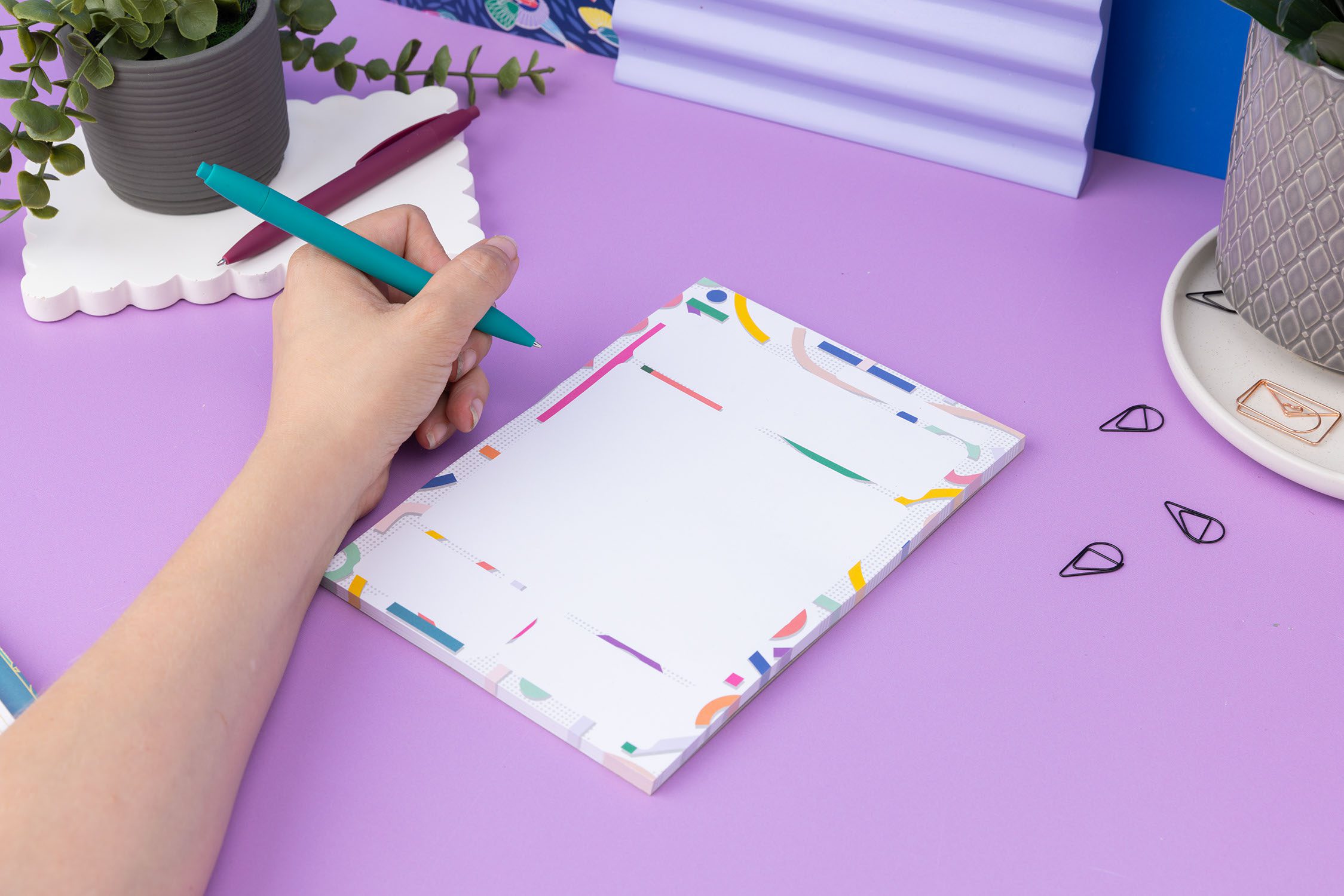 Cutouts A5 Daily Planner Pad on a lilac desk, with a hand to the left holding a teal pen ready to write on it.