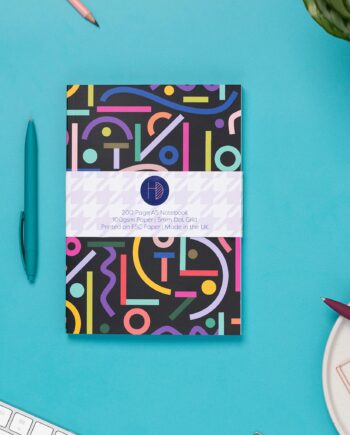 Cutouts A5 Dot Grid Notebook is central, with a branded belly band around it. It is on a teal desk,with pens around it, as well as a white keyboard and a small plant to the top corner.