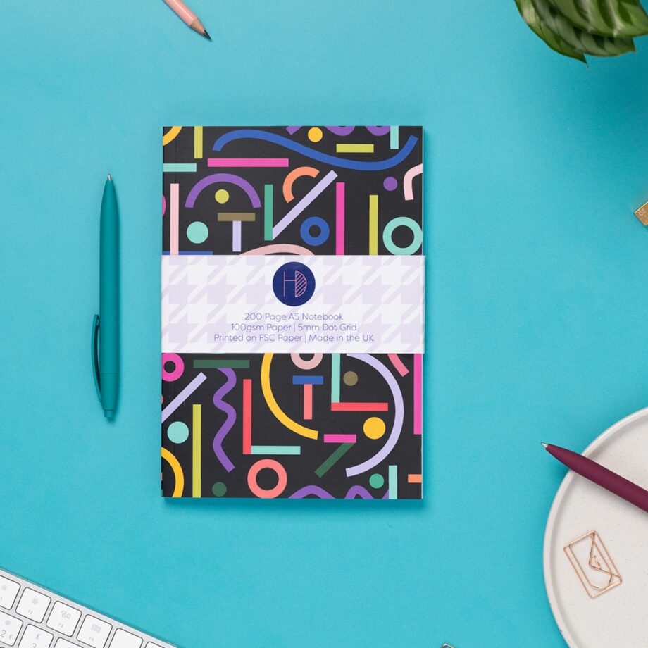 Cutouts A5 Dot Grid Notebook is central, with a branded belly band around it. It is on a teal desk,with pens around it, as well as a white keyboard and a small plant to the top corner.