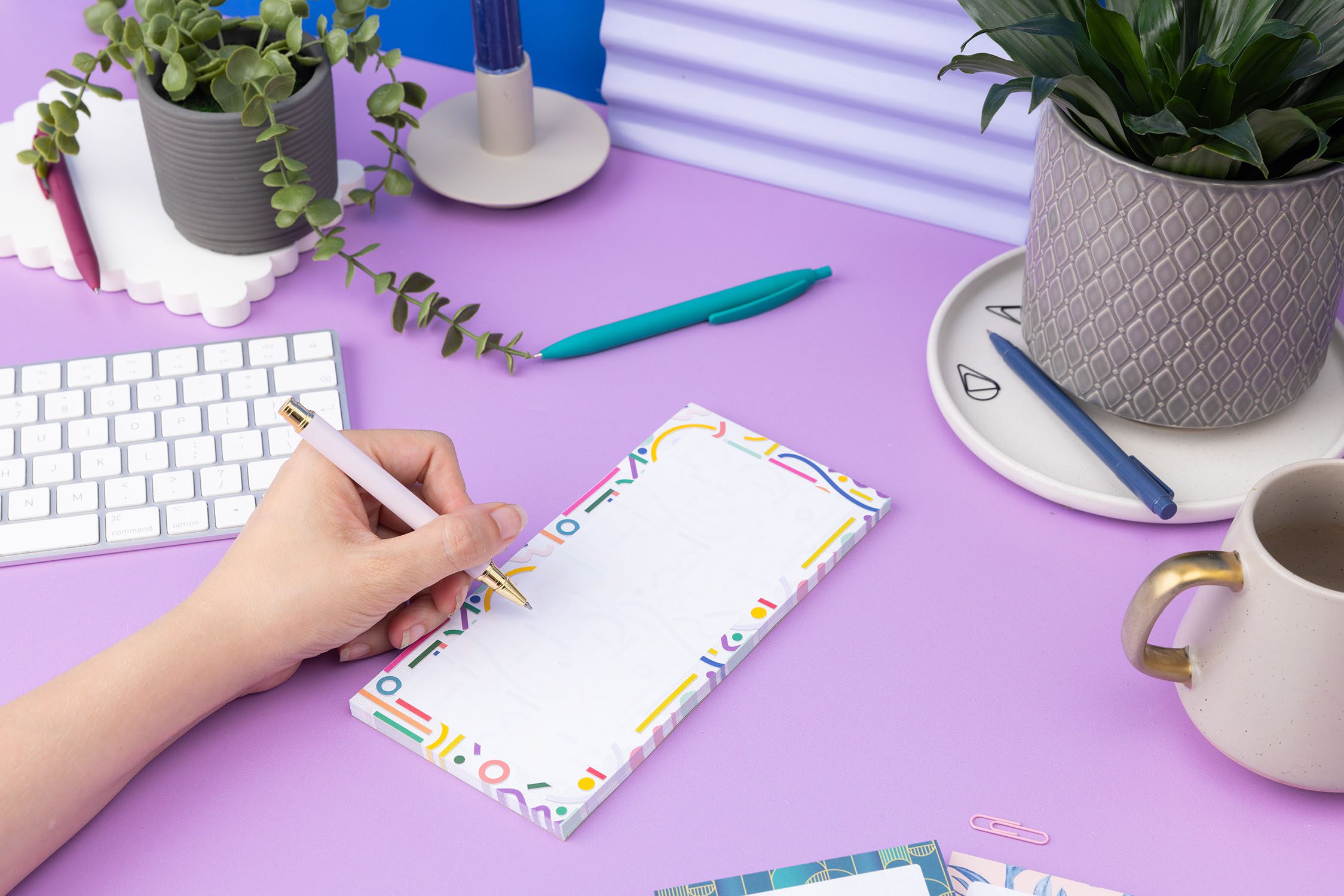 Cutouts List pad is on a lilac desk, with a left hand to its left holding a pen to write on it. There is a small plant, white keyboard and a mug also on the desk around it.