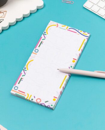Cutouts List pad is on a teal desk, with a white pen leaning on it, with a white keyboard and paperclips around it.