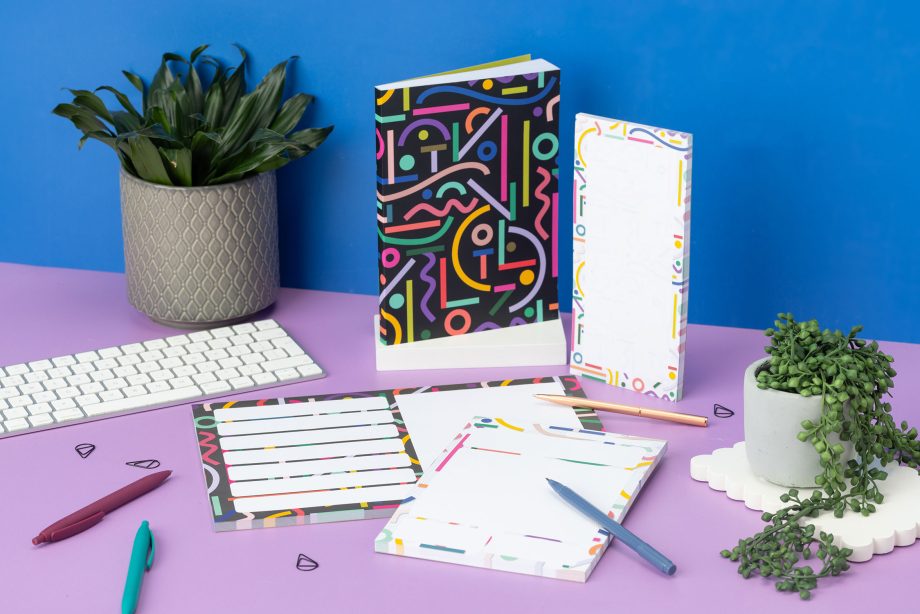 Cutouts Full collection - A4 Planner Pad, A5 Daily Planner Pad, DL List pad and A5 Notebook - are on a lilac desk with small plants, pens and a white keyboard around them. The DL pad is upright, and the A5 Notebook is on a white plinth. There is a blue wall behind.