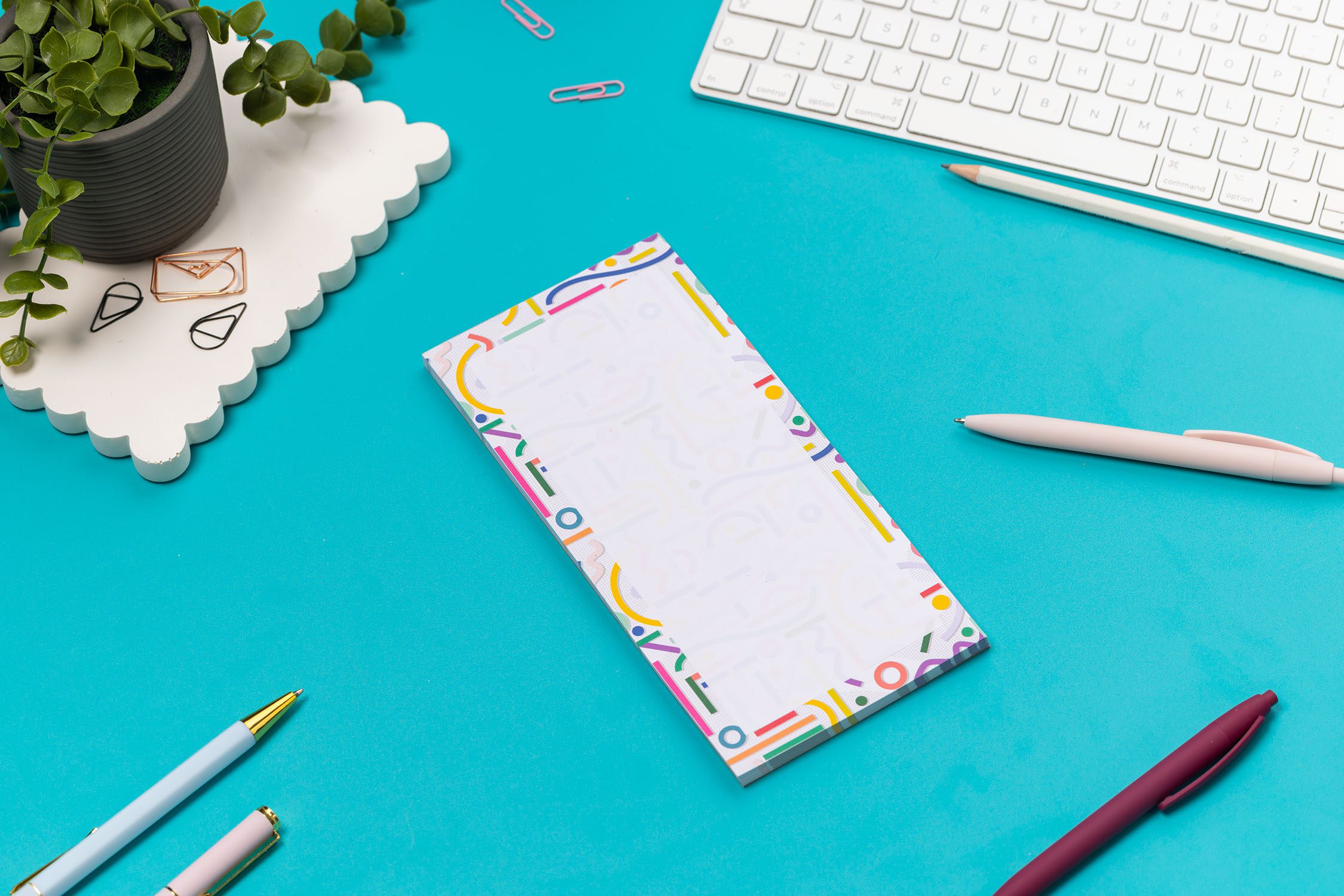 Cutouts List pad is on a teal desk, with a white pen leaning on it, with a white keyboard and paperclips around it.