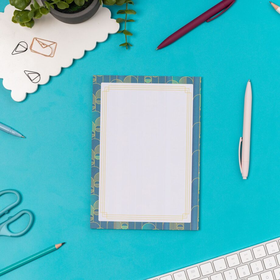 Deco Delights A5 Notepad is on a teal desk with pens and paperclips scattered around it. There is also a white keyboard to the bottom right and small plant to the top left of the image.