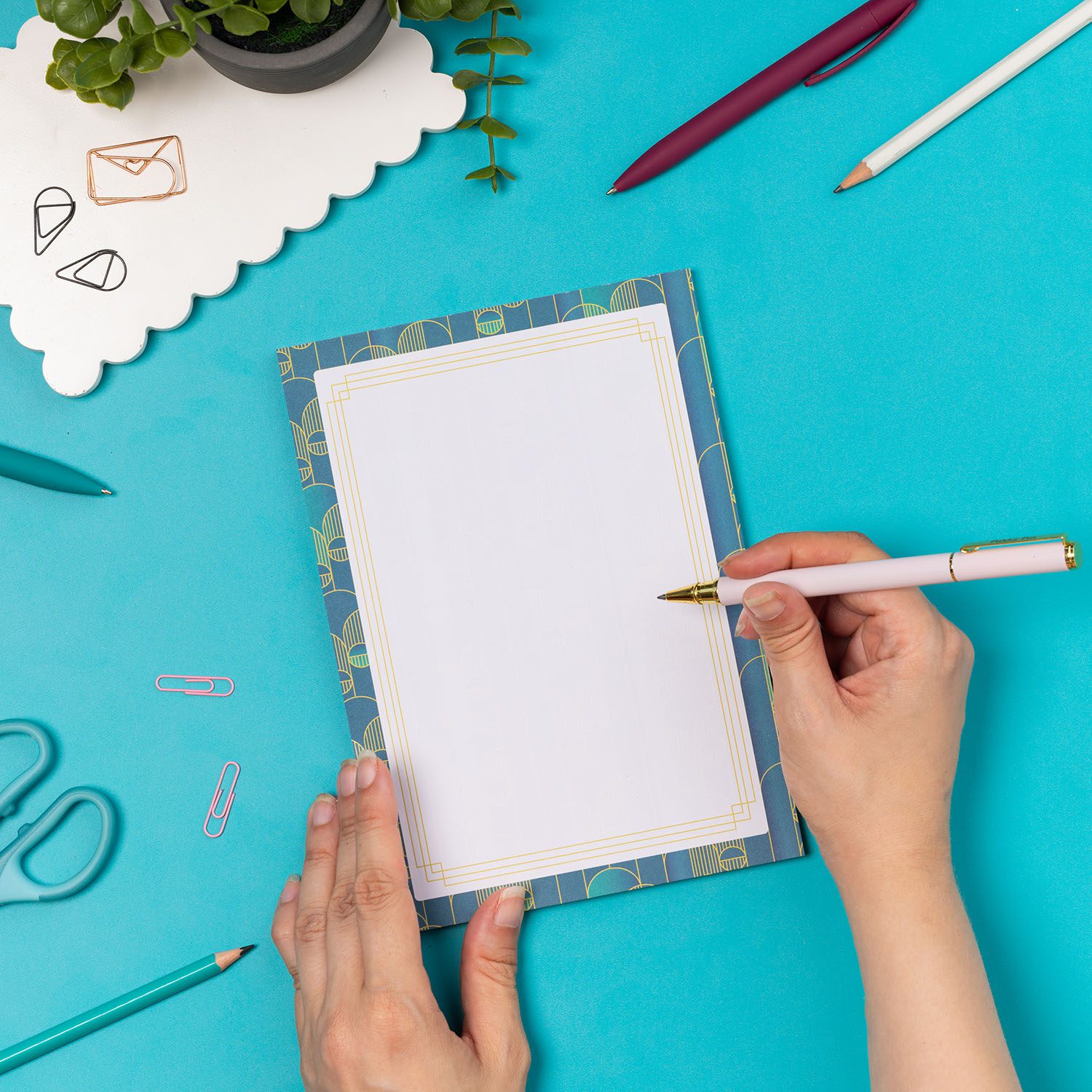 Deco Delights A5 Notepad is on a teal desk with pens and paperclips scattered around it. The pad is being steadied on the desk to its left by one hand, while on its right a hand holding a white and gold pen is about to write on it.