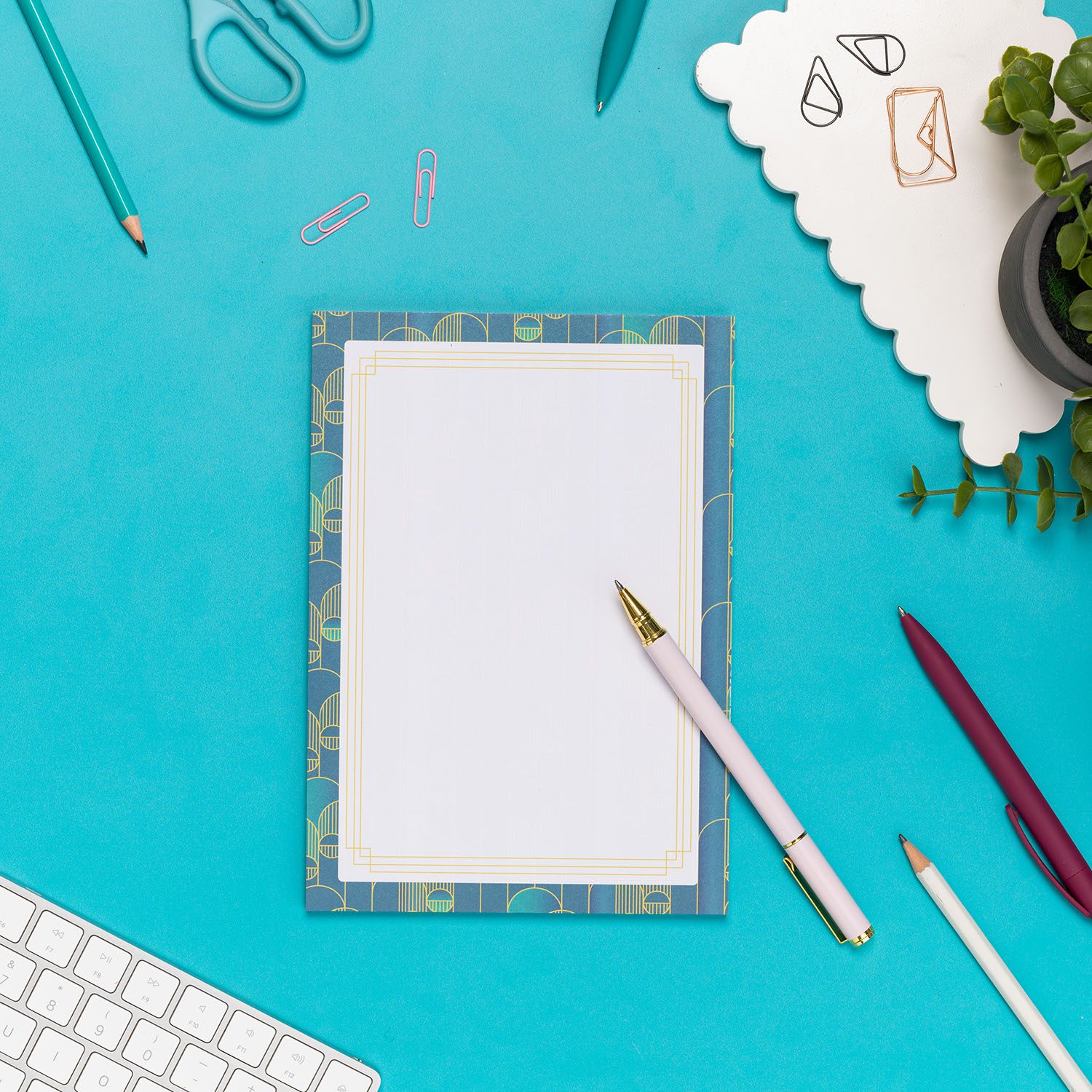 Deco Delights A5 Notepad is on a teal desk with a white pend leaning on it and other pens and paperclips scattered around it. There is also a white keyboard to the bottom left and small plant to the top right of the image.