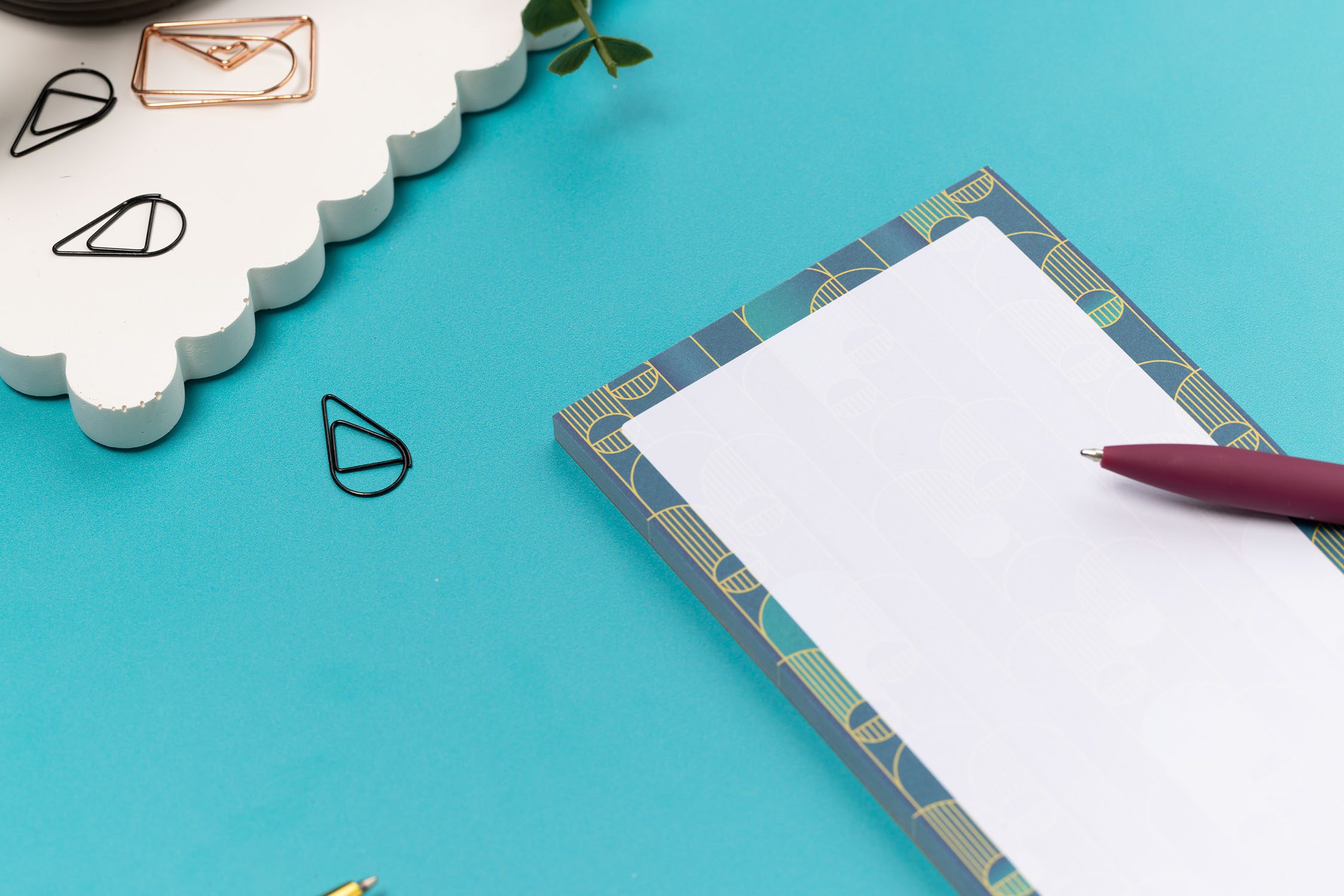 Close up of the top of the Deco delights list pad - teal gradient background with golden yellow deco motif - is on a teal desk with pen and paperclips surrounding it.