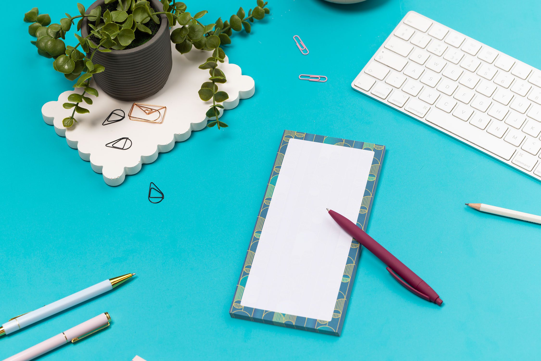 Deco delights list pad - teal gradient background with golden yellow deco motif - is on a teal desk with pen, white keyboard and paperclips surrounding it.