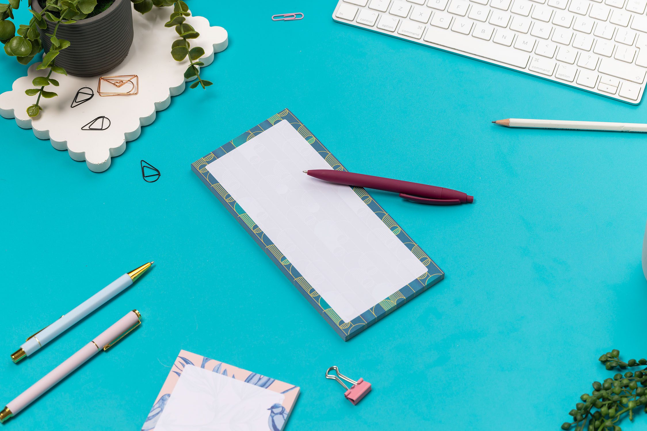 Deco delights list pad - teal gradient background with golden yellow deco motif - is on a teal desk with pen, white keyboard and paperclips surrounding it.