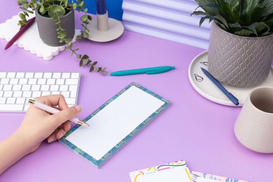 Deco delights list pad - teal gradient background with golden yellow deco motif - is on a lilac desk with pens, pot plants and white keyboard around it. There is a left hand on the left of the image holding a pen to write on the pad.