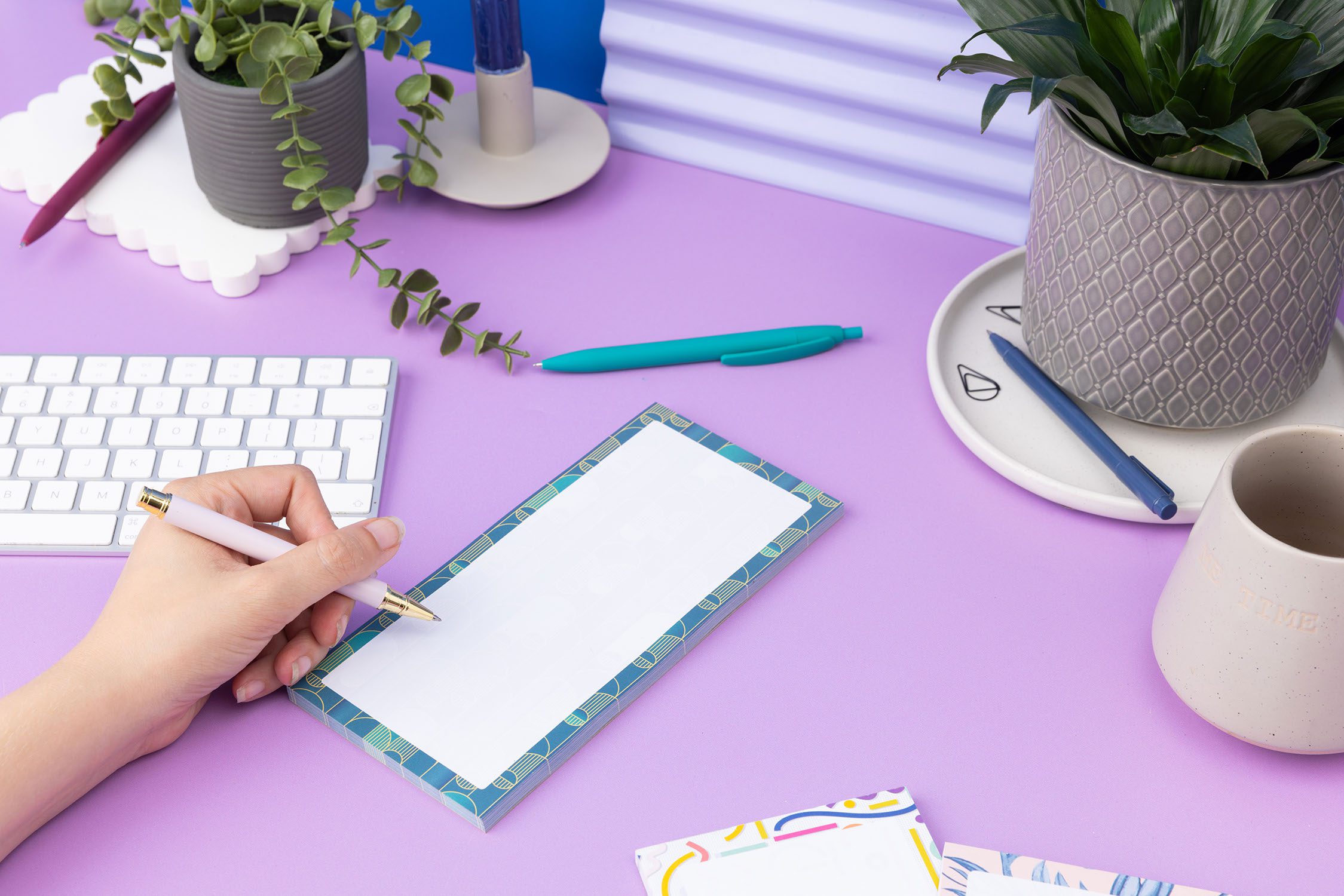 Deco delights list pad - teal gradient background with golden yellow deco motif - is on a lilac desk with pens, pot plants and white keyboard around it. There is a left hand on the left of the image holding a pen to write on the pad.
