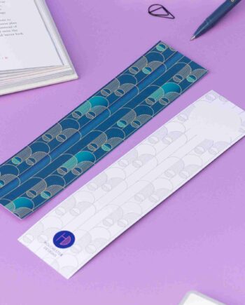 Image of 2 Deco Delights Double sided bookmarks. One with the full-colour side facing up with the teal gradient and golden yellow deco motif. the other has the monochrome version with the deco motif in a light grey and the Holchester Designs logo at the bottom. They are on a lilac desk with an open book to the top right corner.