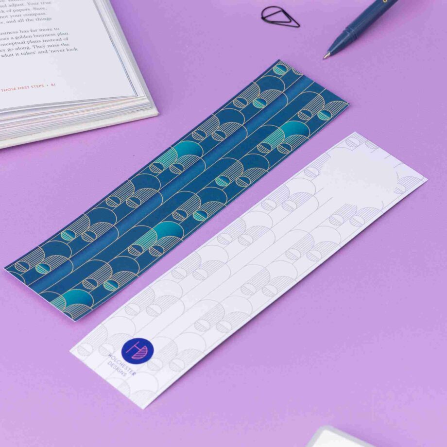 Image of 2 Deco Delights Double sided bookmarks. One with the full-colour side facing up with the teal gradient and golden yellow deco motif. the other has the monochrome version with the deco motif in a light grey and the Holchester Designs logo at the bottom. They are on a lilac desk with an open book to the top right corner.