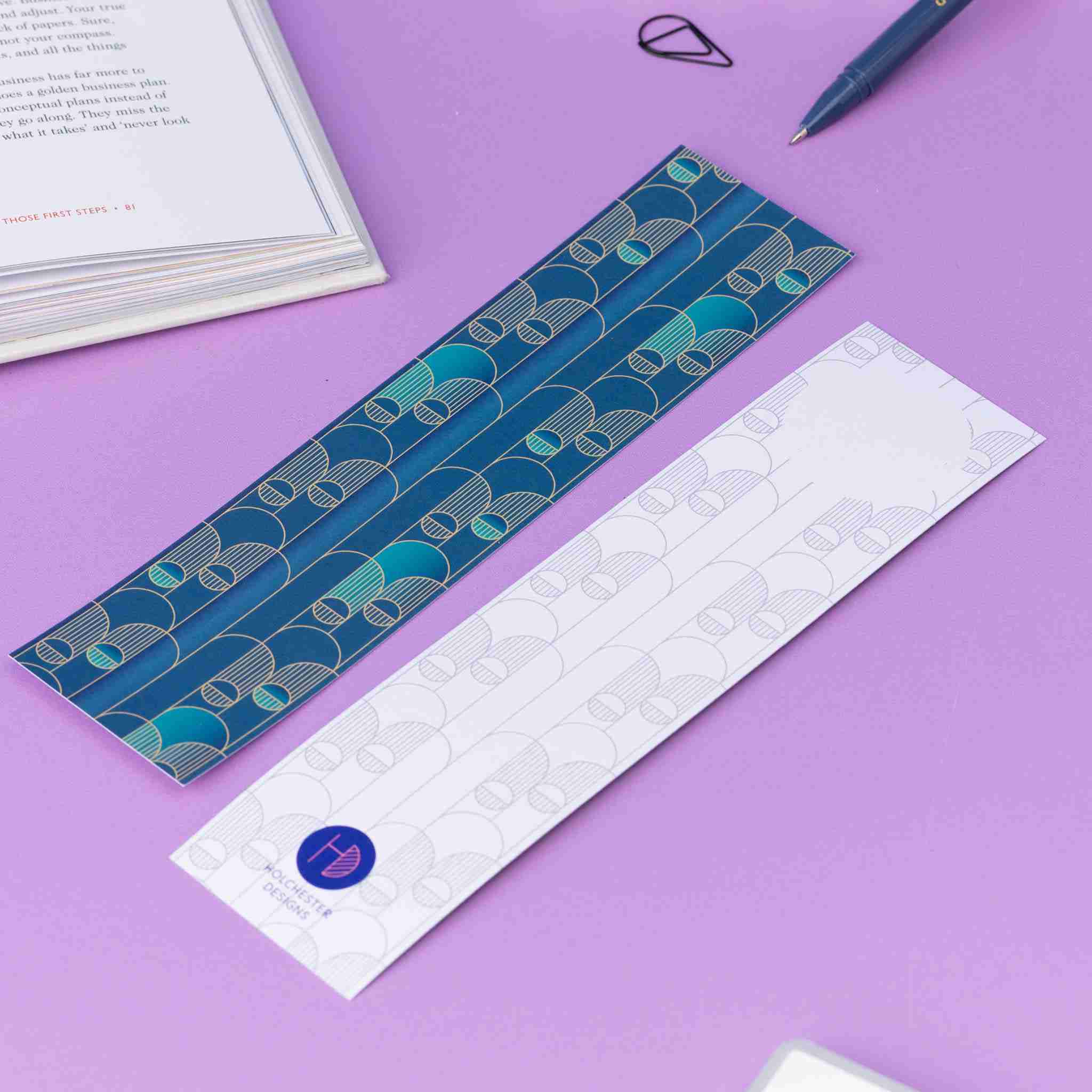 Image of 2 Deco Delights Double sided bookmarks. One with the full-colour side facing up with the teal gradient and golden yellow deco motif. the other has the monochrome version with the deco motif in a light grey and the Holchester Designs logo at the bottom. They are on a lilac desk with an open book to the top right corner.