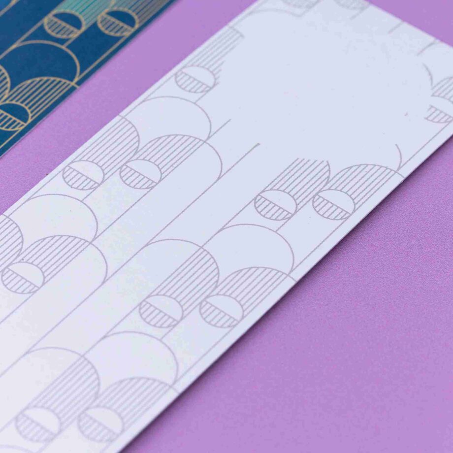 Image of 2 Deco Delights Double sided bookmarks. The focus is on the top of the monochrome reverse side, showing the blank circle for writing in. Next to it is a full-colour side up, out of focus to the left. They are lying on a lilac desk.