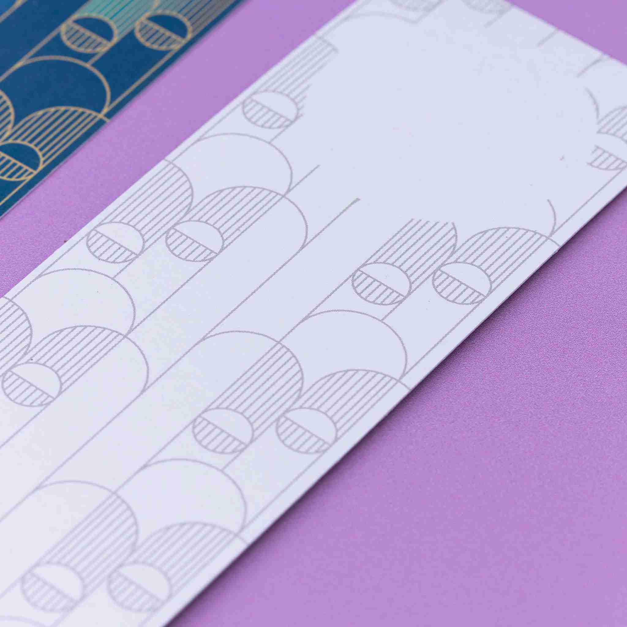 Image of 2 Deco Delights Double sided bookmarks. The focus is on the top of the monochrome reverse side, showing the blank circle for writing in. Next to it is a full-colour side up, out of focus to the left. They are lying on a lilac desk.
