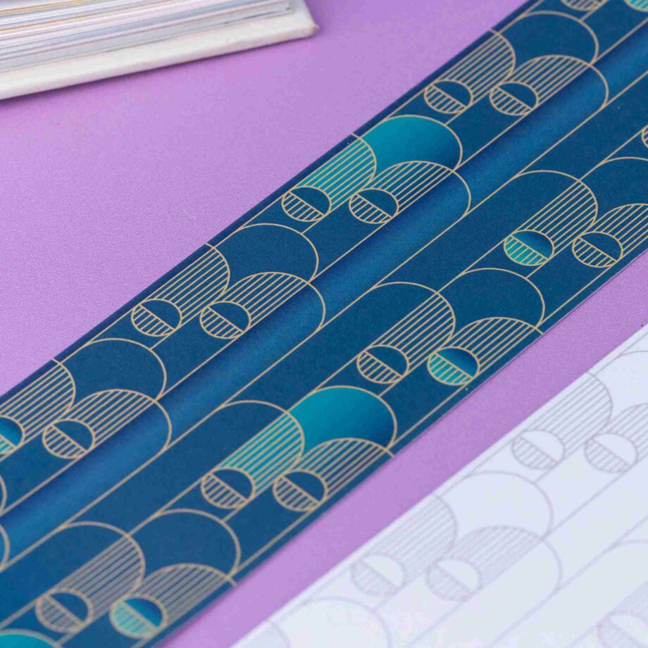 Close up of the full-colour patterned side of the Deco Delights bookmark with the teal gradient background and the golden yellow repeat motif. Next to it is an out of focus corner of the bookmark's other side. They are on a lilac desk with an open book in the top left corner.