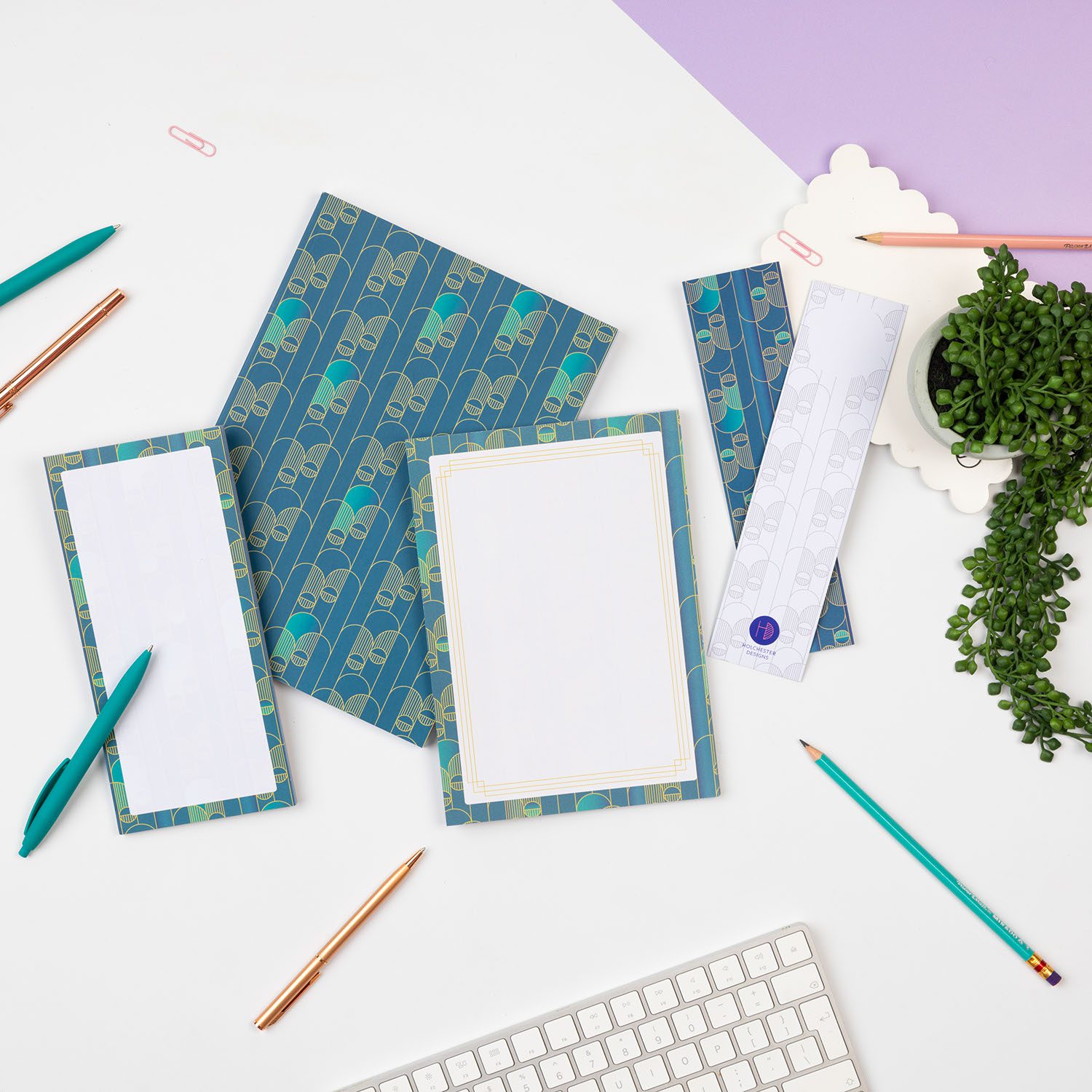 Deco Delights Full collection are laid in a row across a white desk with pens scattered around them and a plant to the right. From left to right there is the List Pad, B5 Dot grid notebook, A5 notepad and two double-sided bookmarks (one of each side facing up).