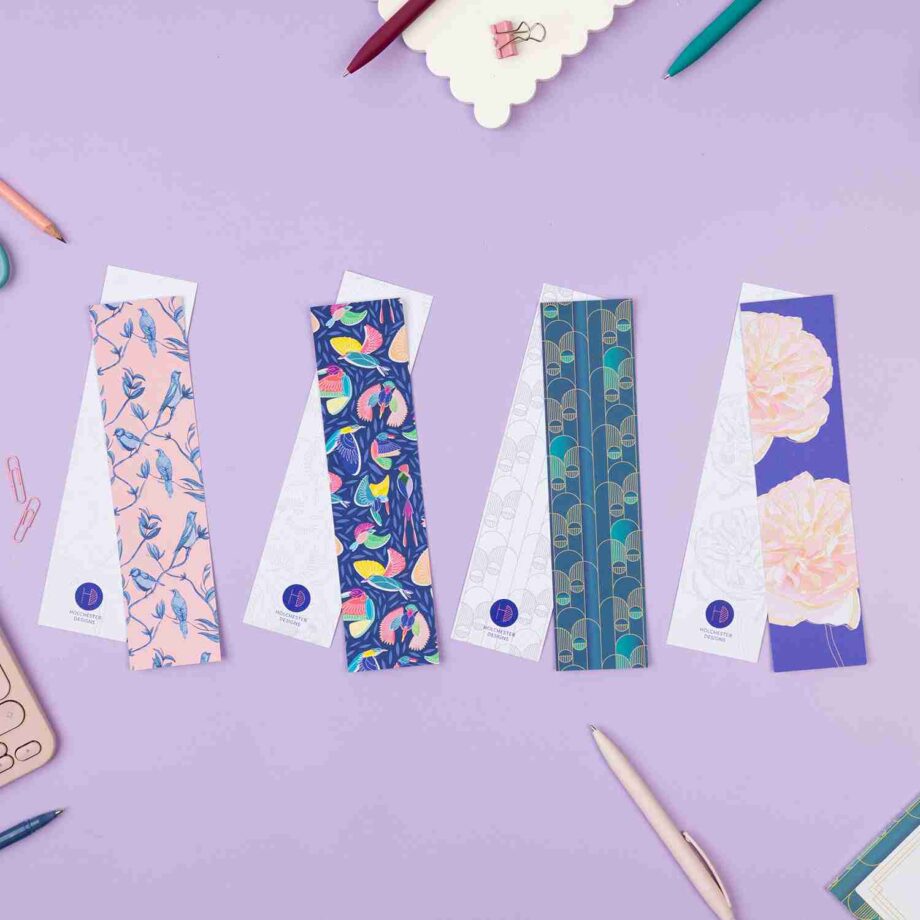 4 pairs of bookmarks are in a row on a lilac table with pens and pencils around them. From Left to right there are Brighton Birds, Paradise, Deco Delights and Spring Forward. Each have the full-colour version face up on top of the monochrome sides underneath.