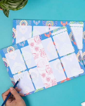 An A4 Planner Pad, featuring the metamorphosis pattern, has a white belly band around it with a pink floralline design on it. It is lyingon an angle on top of another identical planner pad, with a hand to its left holding a pencil.