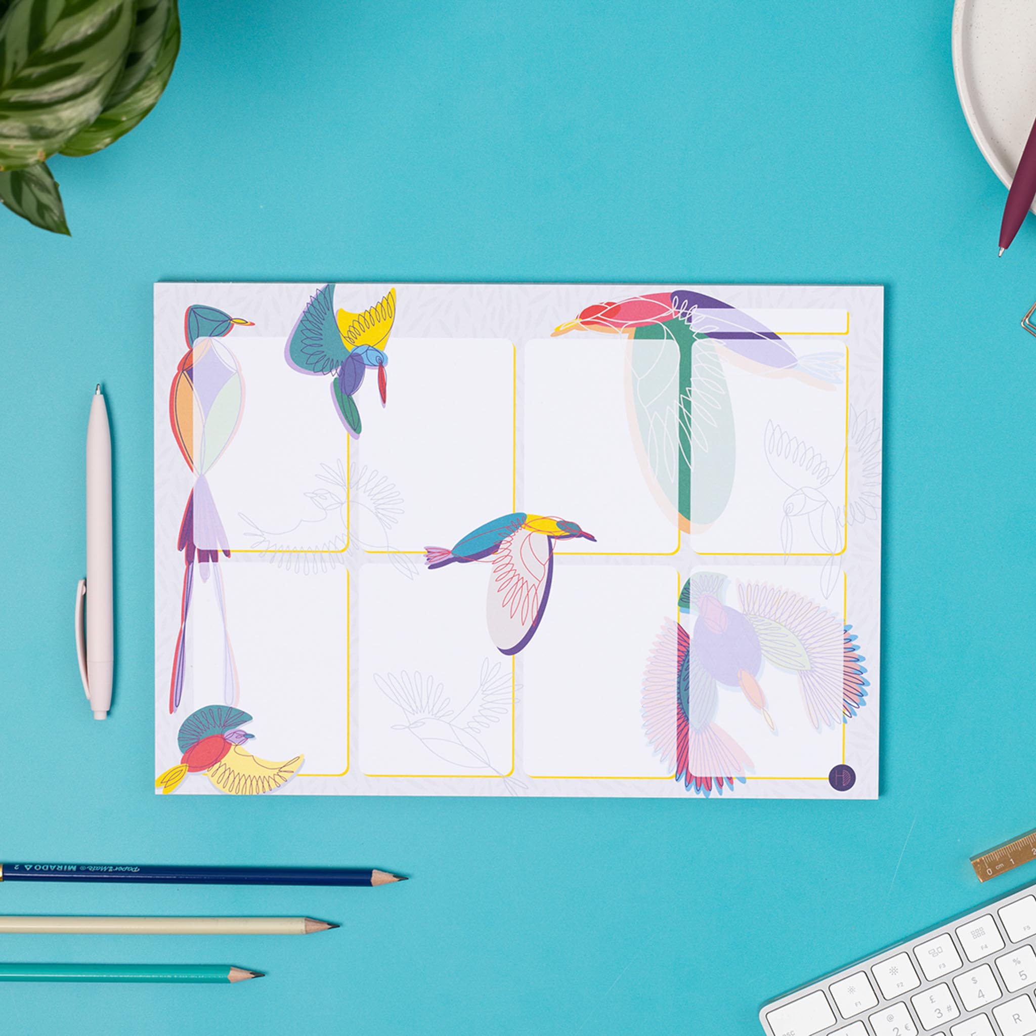 A4 Planner pad with a light grey background and various multi-coloured line drawn bird illustrations around it. there are 8 white boxes, with a top row for a date, and the birds are mainly behind these, although some overlap at the edges. The pad is on a teal desk with a white keyboard and pens surrounding it.