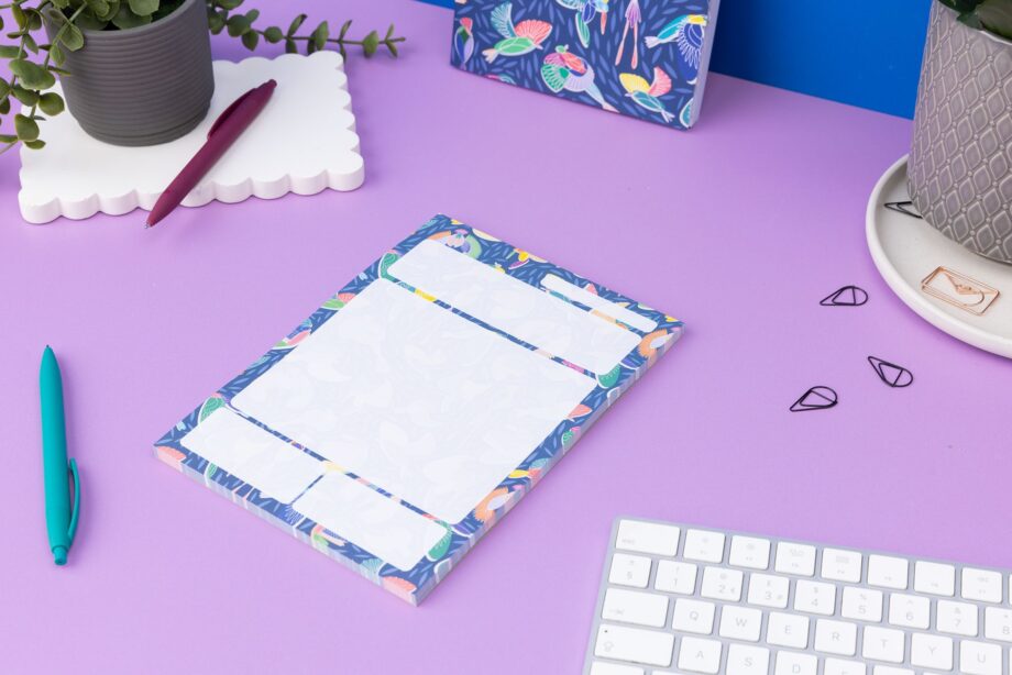Paradise A5 Daily Planner Pad is central on a lilac desk, with pens, paperclips and a white keyboard around it. To the top you can see a pot plant and a Paradise Dot Grid Notebook leaning against a blue wall.