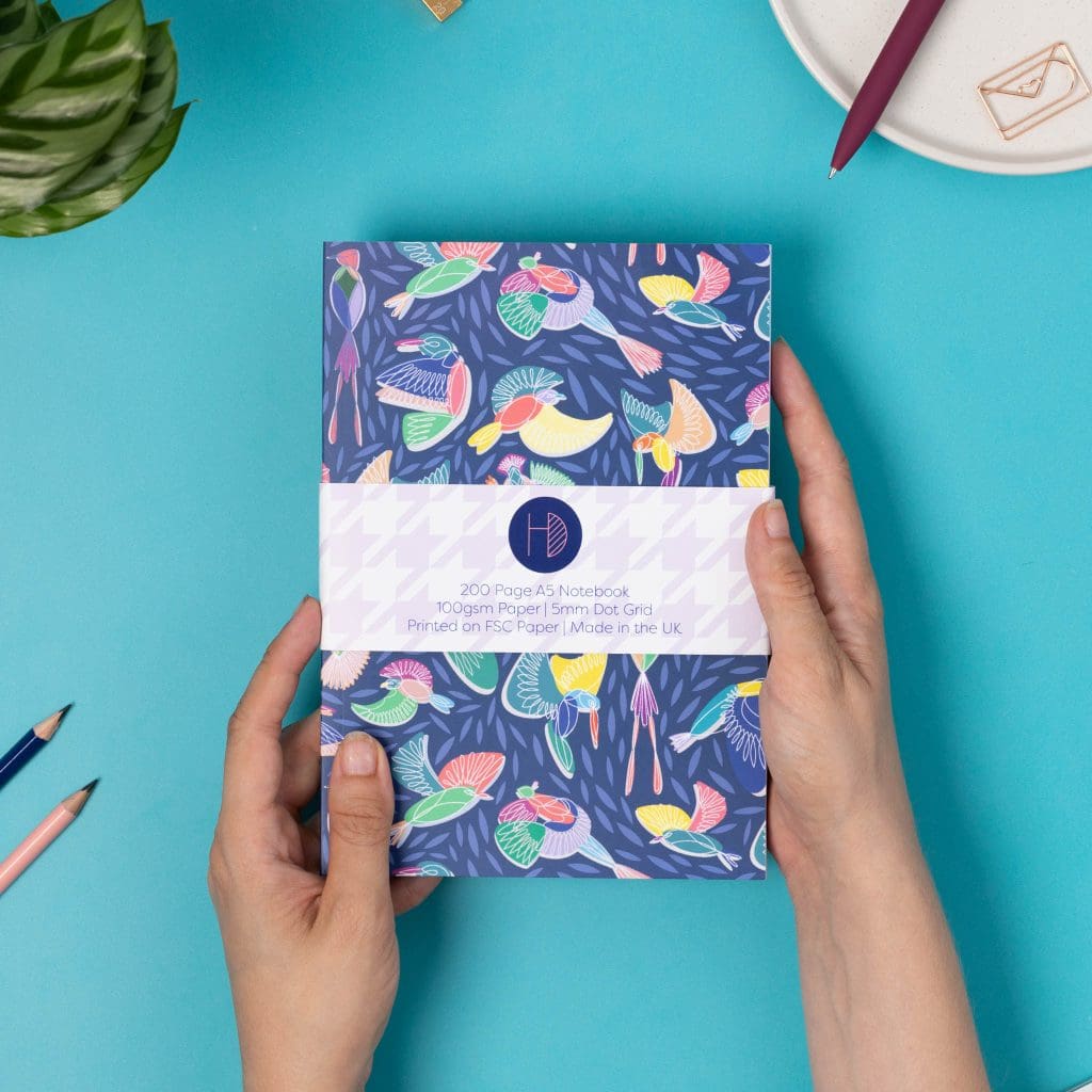 Two hands hold either side of a Paradise A5 Dot Grid Notebook, with its belly-band packaging around it, is on a teal desk with some pens, and a keyboard to the bottom left corner. The book's cover features a navy background with line-drawn bird motifs and offset coloured blocks behind them.