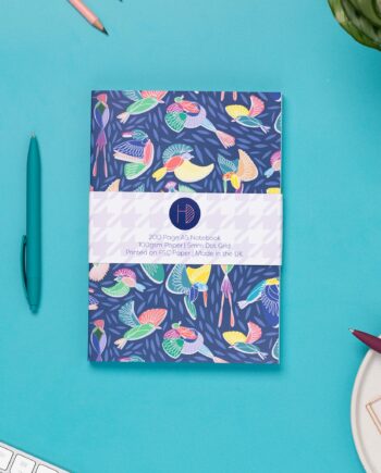 Paradise A5 Dot Grid Notebook, with its belly-band packaging around it, is on a teal desk with some pens, and a keyboard to the bottom left corner. The book's cover features a navy background with line-drawn bird motifs and offset coloured blocks behind them.