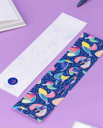 Two of the paradise double-sided bookmarks, on a lilac desk with a book open to the top left corner. One side features the full-colour patterned side of the Paradise pattern with the navy background, bird line motifs and colour blocks behind. The other side is the light-grey line drawing.