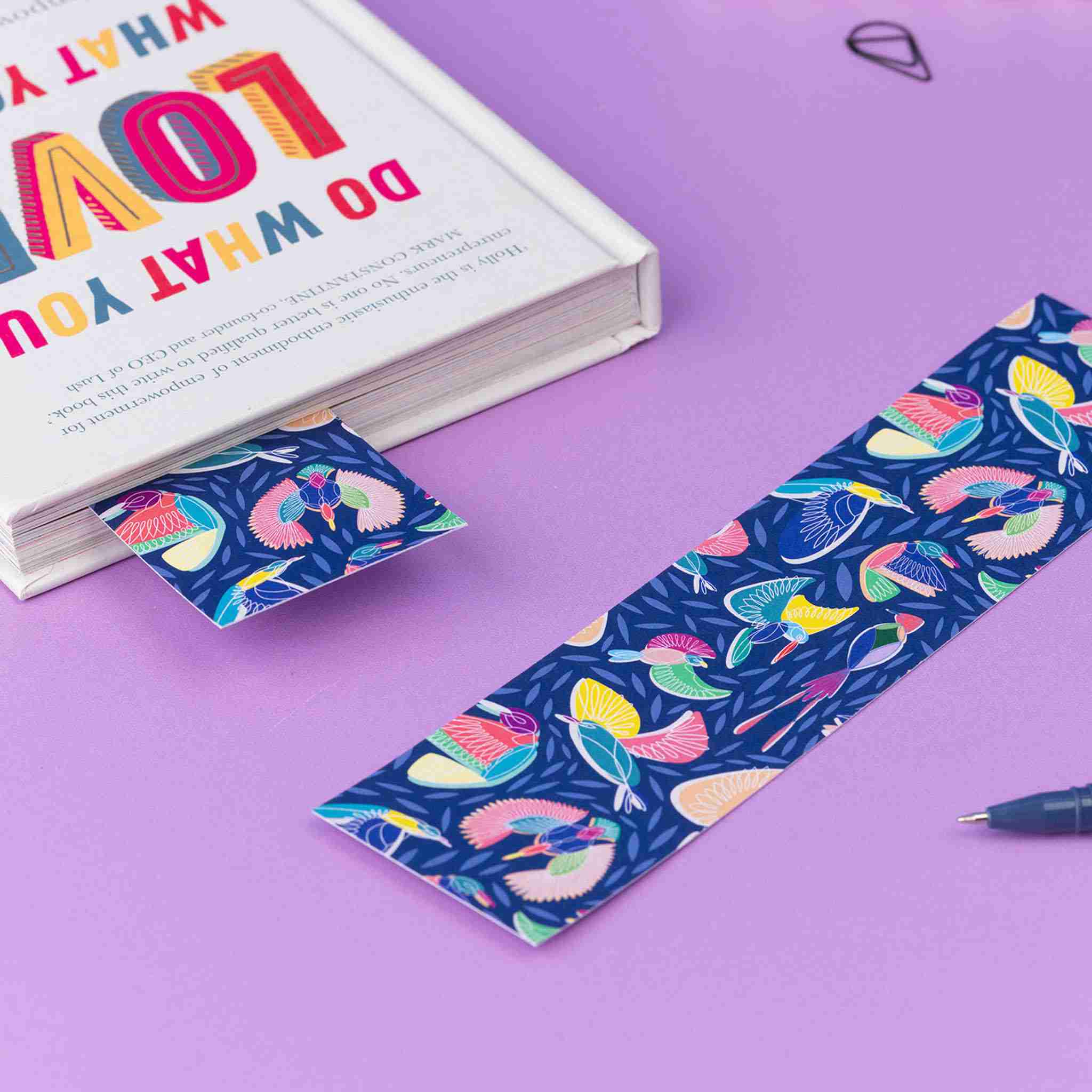 A Paradise double-sided bookmark is lying on a lilac desk with its full-colour side facing up. To its left is a close book which has a Paradise bookmark sticking out of it, also full-colour side up.