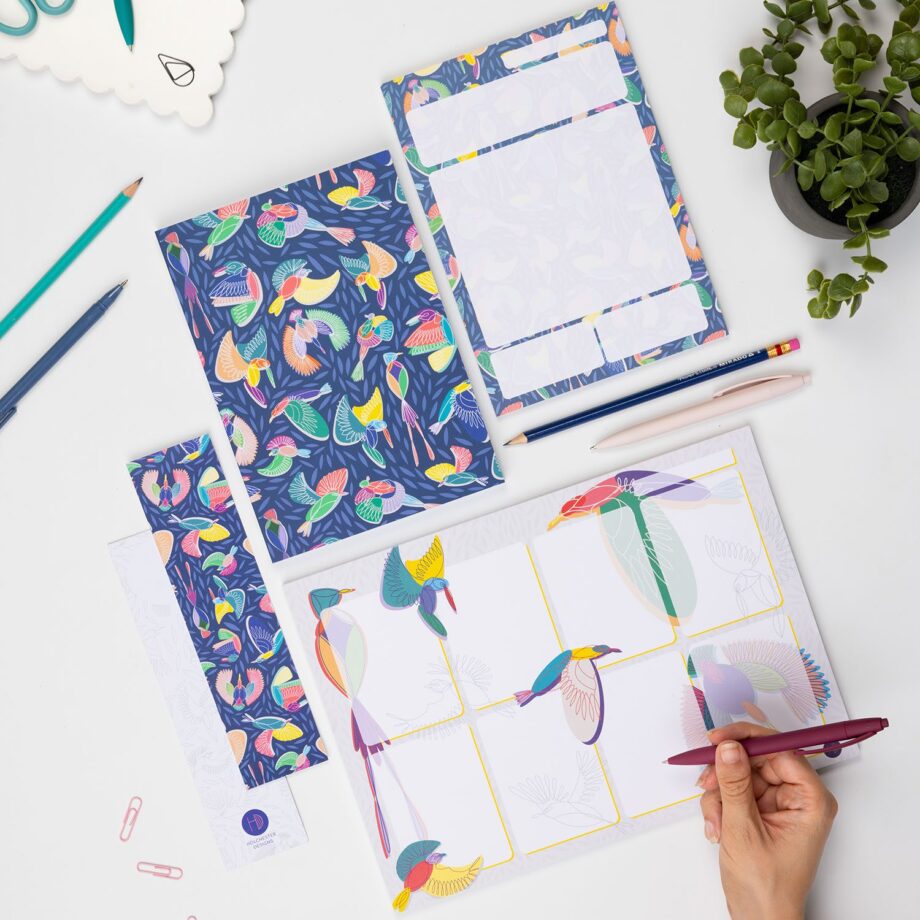 The Full Paradise collection - A4 Planner pad, A5 notebook, A5 Daily Planner Pad and 2 Double-side bookmarks (one side each) - are lying on a white desk surrounded by pens, paperclips and a small plant. There is a right hand in the bottom right corner holding a pen to write on the planner pad.